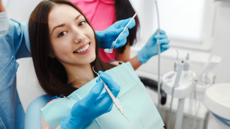 Junge lächelnde Frau im Zahnarztstuhl. Kieferorthopäde mit Handschuhen hält Mundspiegel und Explorer für die Untersuchung. Zahnarzthelferin im Hintergrund mit Sauger.